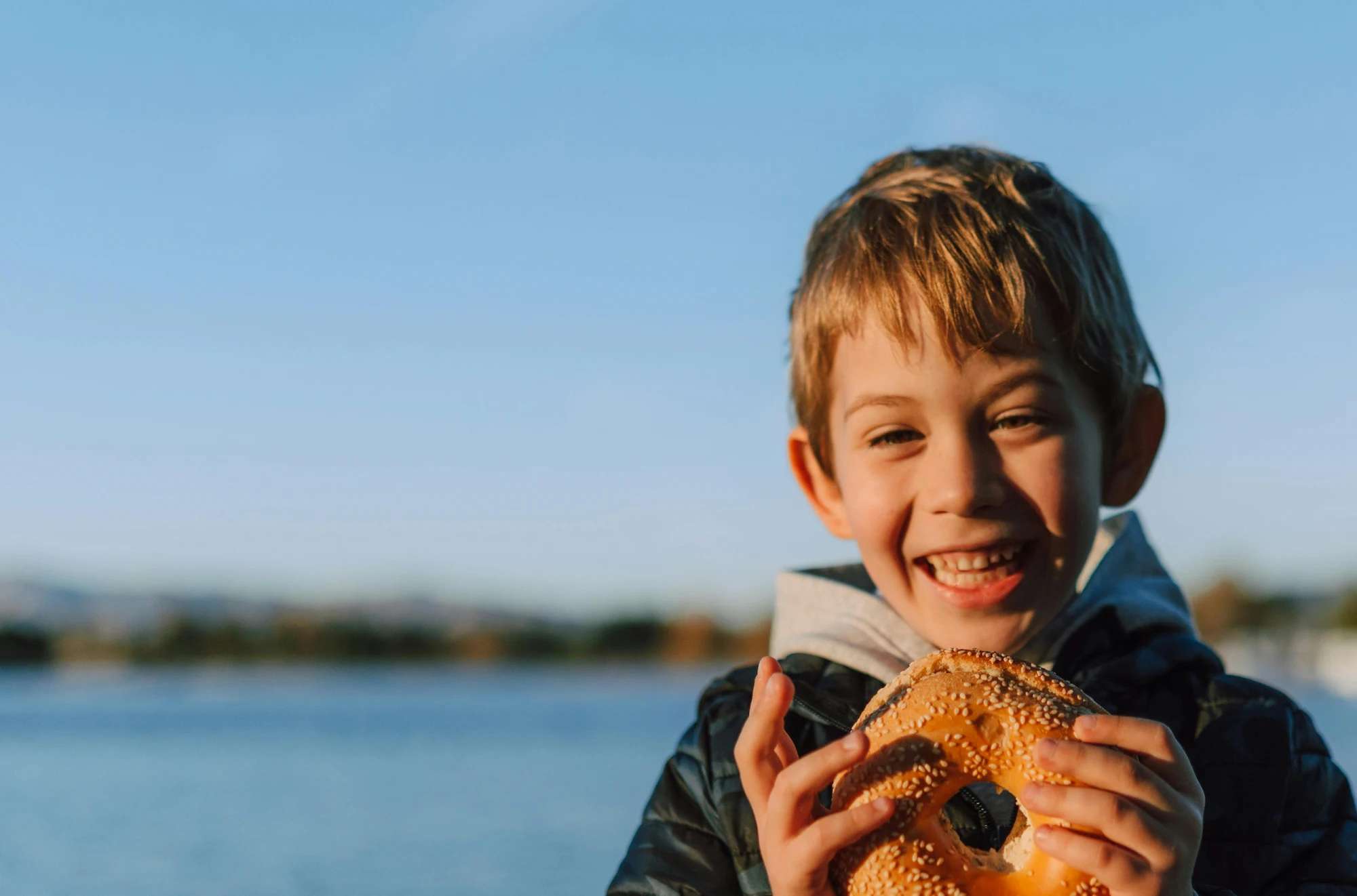 Young boy smiling with a bagel by a body of water